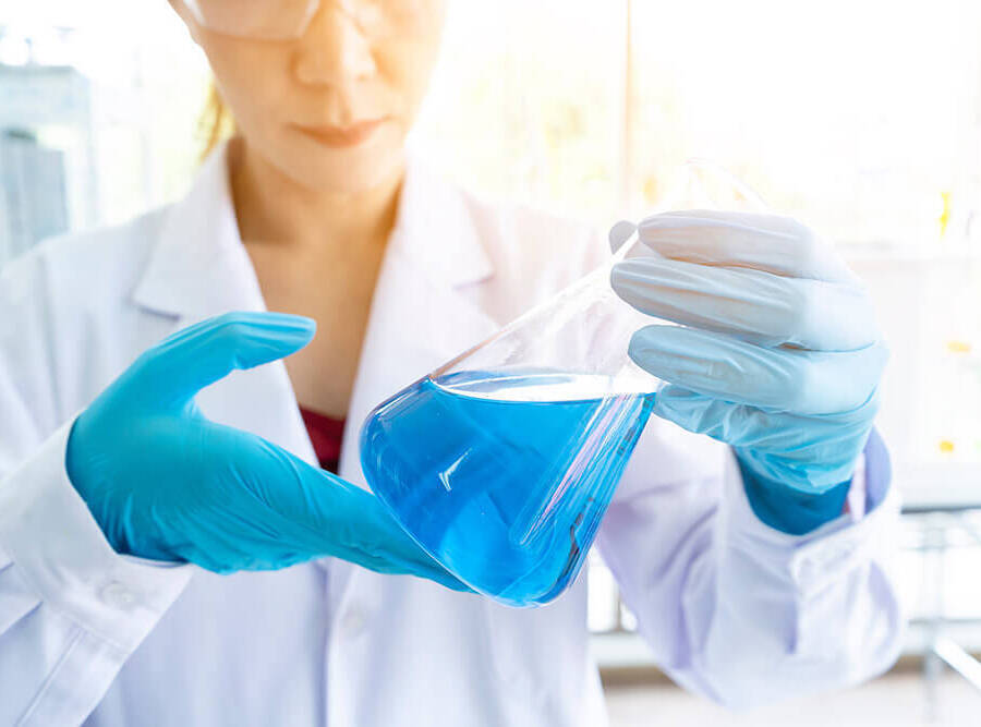 Technician in lab coat and blue rubber gloves, looking at and tilting a conical flask filled with blue liquid