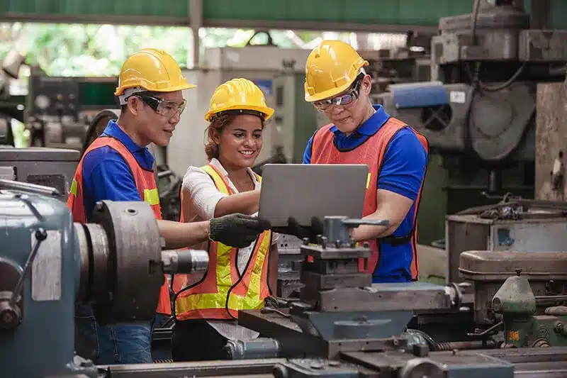 Three workers in hard hats and high visibility vests huddled looking at a laptop, lathes and manufacturing equipment surrounds them