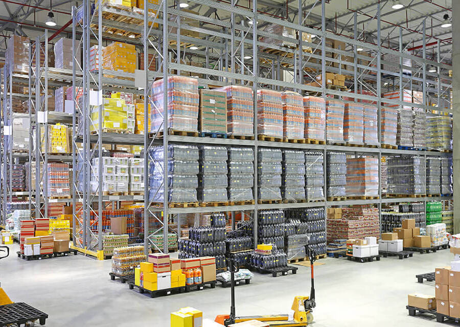A fully stocked warehouse, with shelves filled with pallets to the ceiling, in the foreground are more pallets of boxes and pallet trucks.