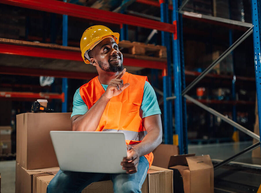 Warehouse employee wearing safety gear, sat on boxes, pointing to the sky in a warehouse setting